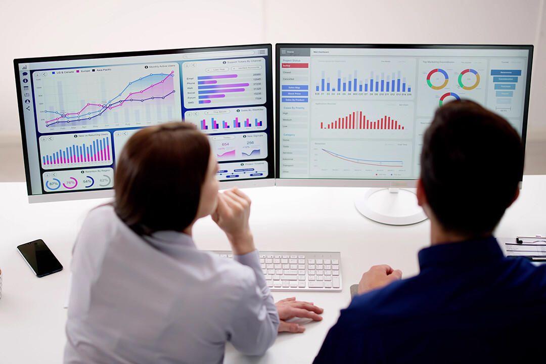 Two people reviewing marketing analytics dashboards with performance graphs, audience data, and conversion metrics on dual monitors.