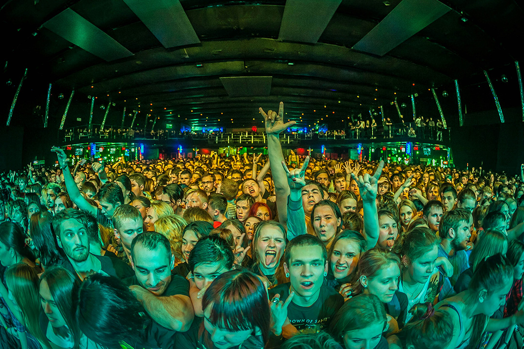 A large crowd of fans gather in the front rows of a concert stage with green and orange lights spotlighting the larger audience.