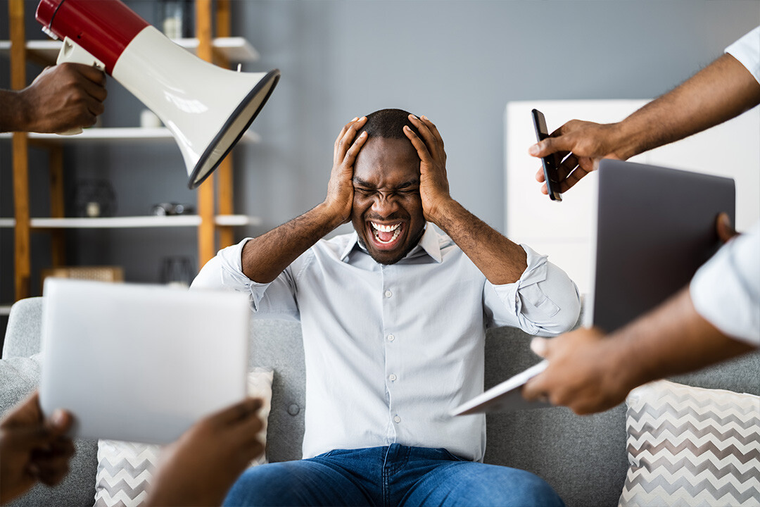 A frustrated man sitting on a couch, holding his head in distress, while multiple people overwhelm him with documents, a laptop, a phone, and a megaphone. This image represents customer frustration due to ad overload, poor targeting, and ineffective engagement strategies, which can lead to high churn rates.