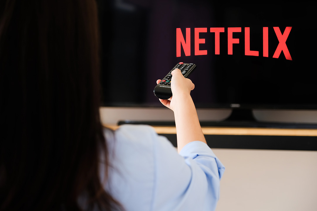 A woman watching television, holding a remote control with the Netflix streaming home screen in the background.
