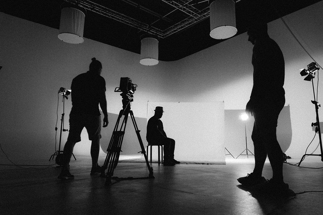 A black and white image of a camera crew filming actors, lighting setup, and candid moments between takes.