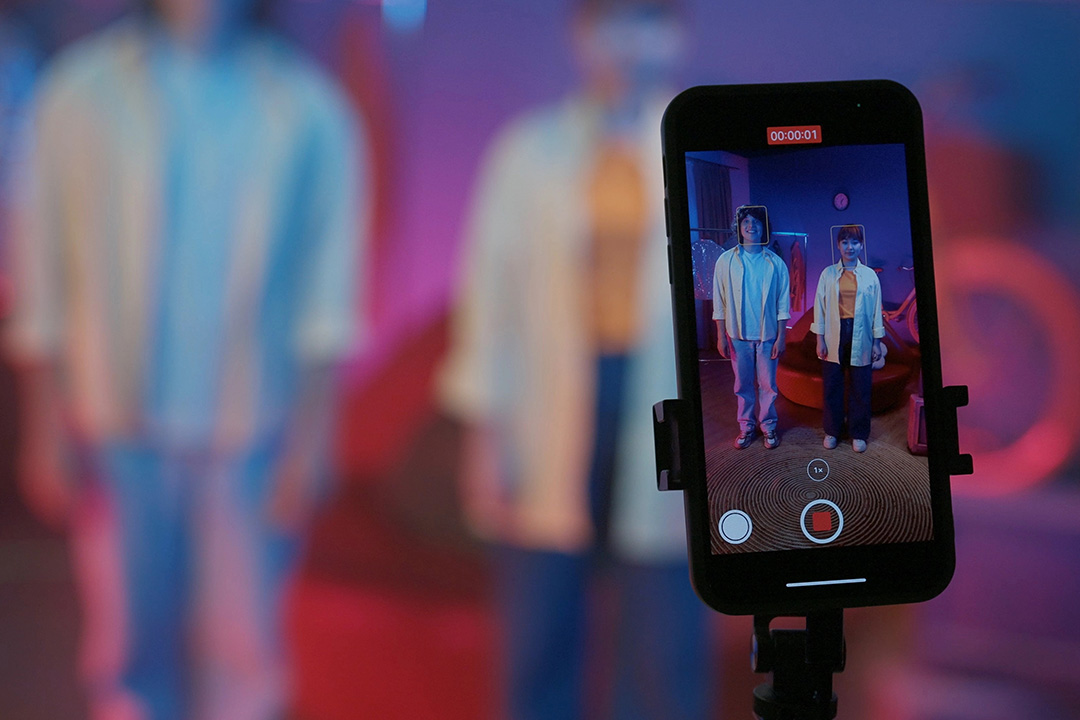 Smartphone screen showing two young content creators recording a social media video, with vibrant lighting and a blurred background suggesting a modern digital production setup.
