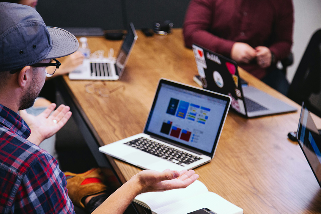 A diverse group of creative professionals collaborating around a table with laptops, notebooks, and coffee cups, discussing marketing strategies. They are engaged in brainstorming ideas related to the customer journey, with visuals of journey maps or strategy notes visible on their screens. The atmosphere appears dynamic and focused, reflecting a team working together to develop innovative marketing solutions