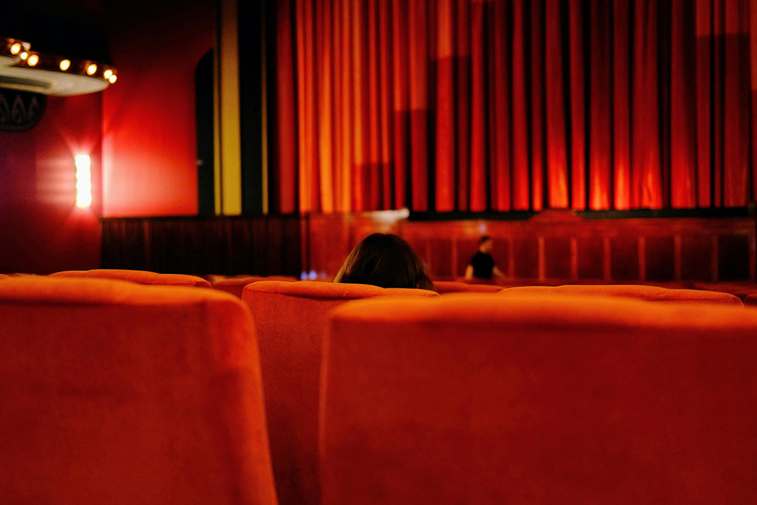 An image of a traditional old-school movie theater with a red curtain covering the screen, red plush cinema seats; representing film distribution.